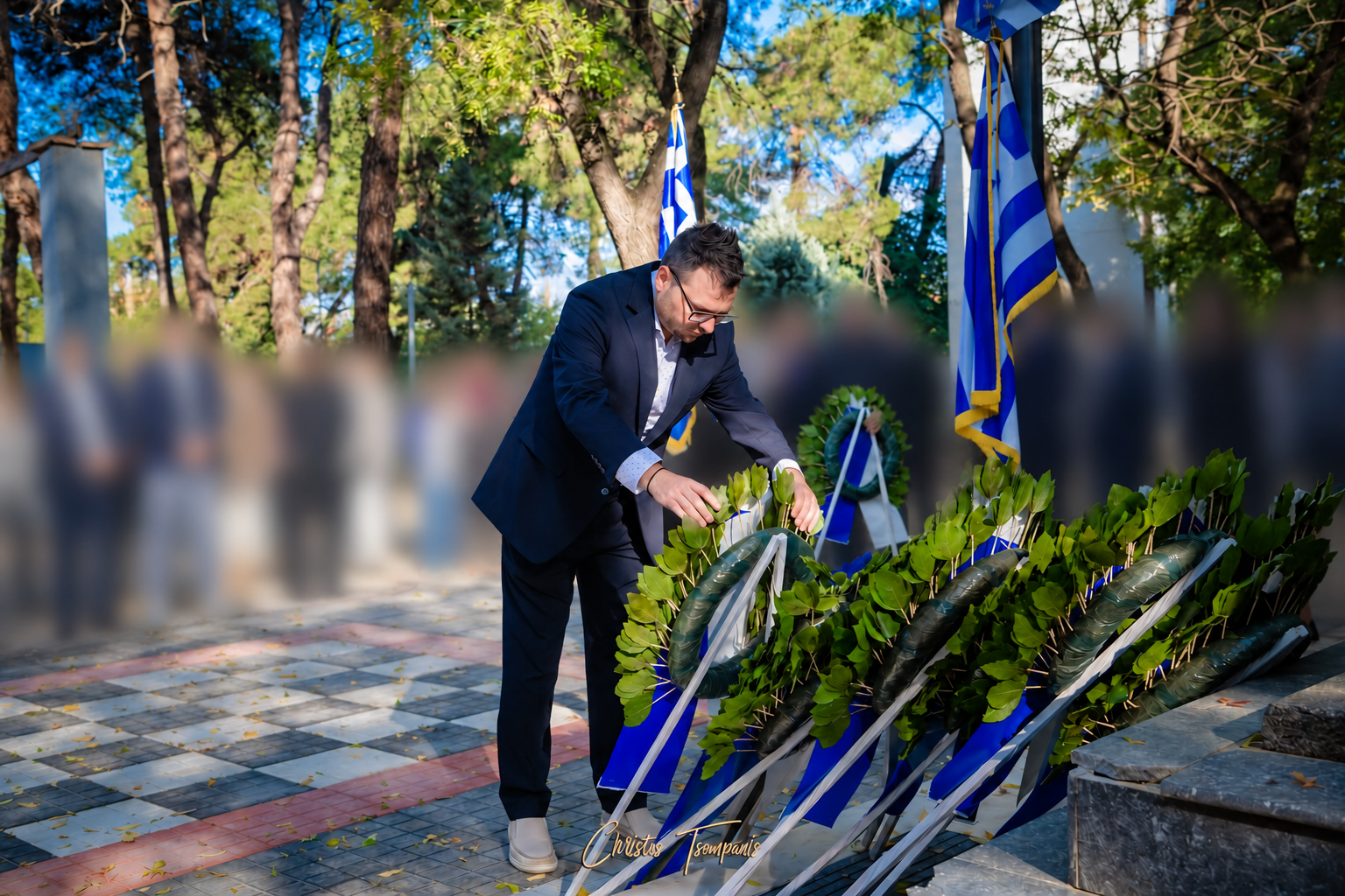 Ο Αντώνης Μάνος καταθέτει στεφάνι, με θολωμένους παρευρισκόμενους στο φόντο.